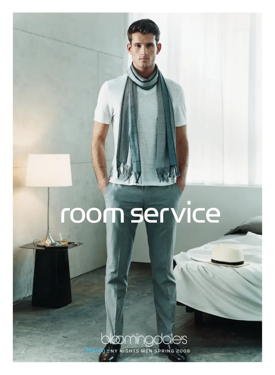 A male model wearing a Theory white t-shirt and scarf, standing in a hotel room setting. Campaign by PlanetFab Studio.