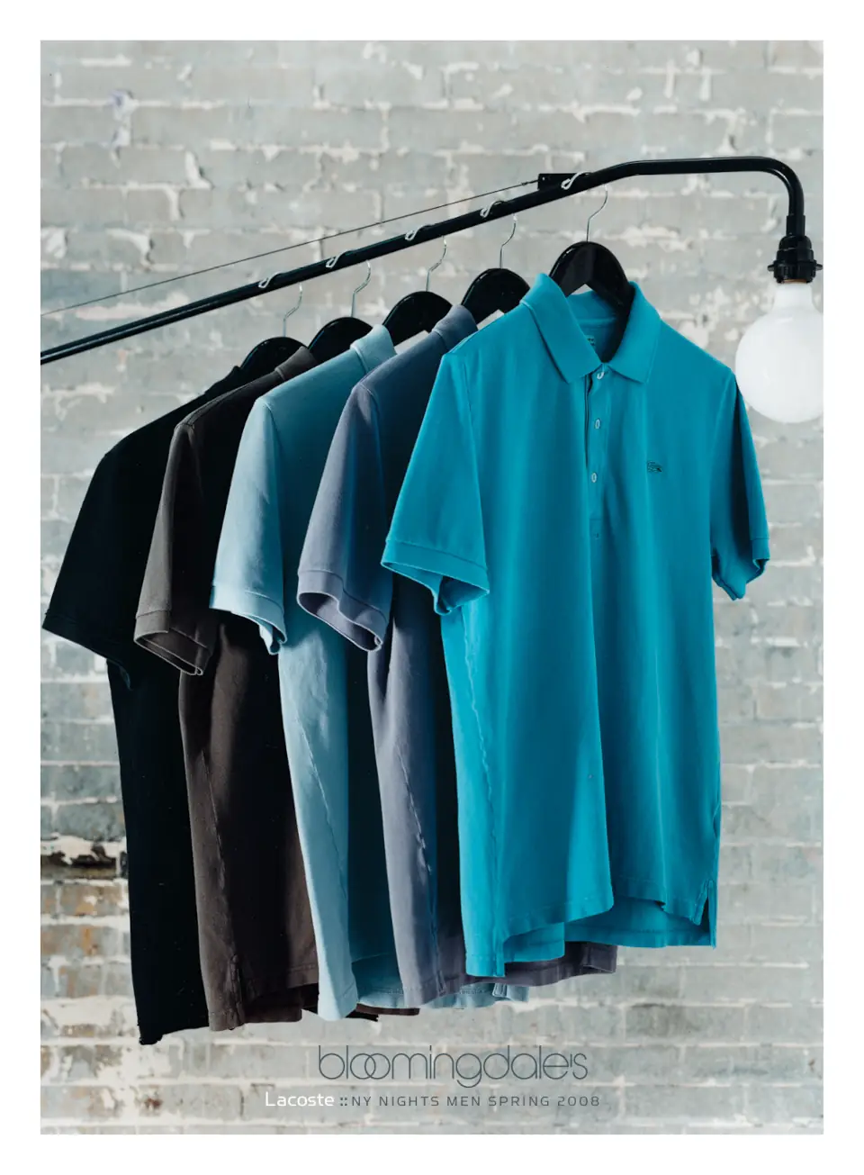 Blue and grey Lacoste polo shirts on hanging on a lamp displayed in front of a brick wall. Campaign by PlanetFab Studio.