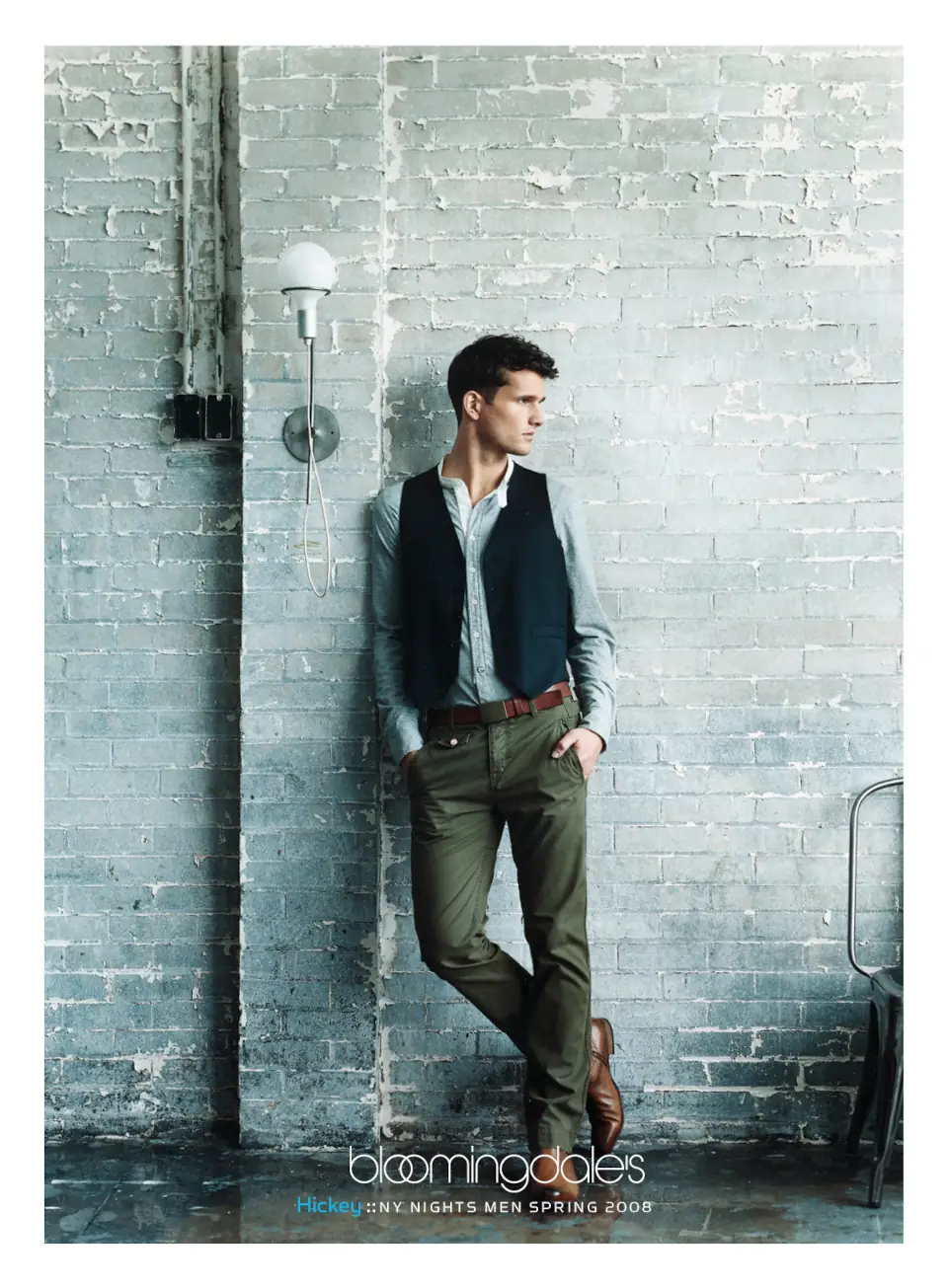 A male model wearing a Hickey Freeman vest and olive pants, leaning against a painted brick wall. Campaign by PlanetFab Studio.