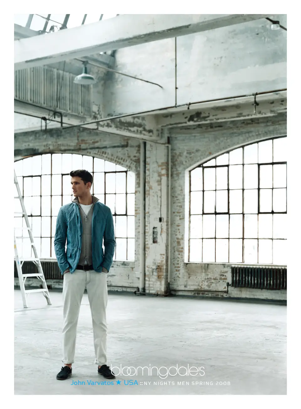 A male model wearing a John Varvatos teal jacket standing in an industrial space with large windows. Campaign by PlanetFab Studio.