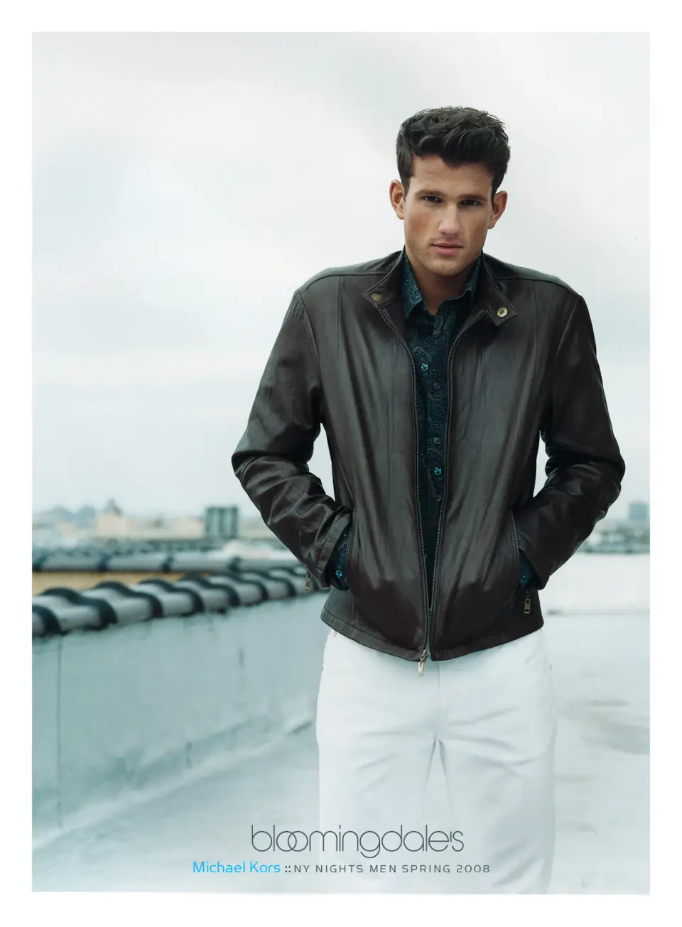 A male model wearing a Michael Kors dark leather jacket and white pants on a rooftop. Campaign by PlanetFab Studio